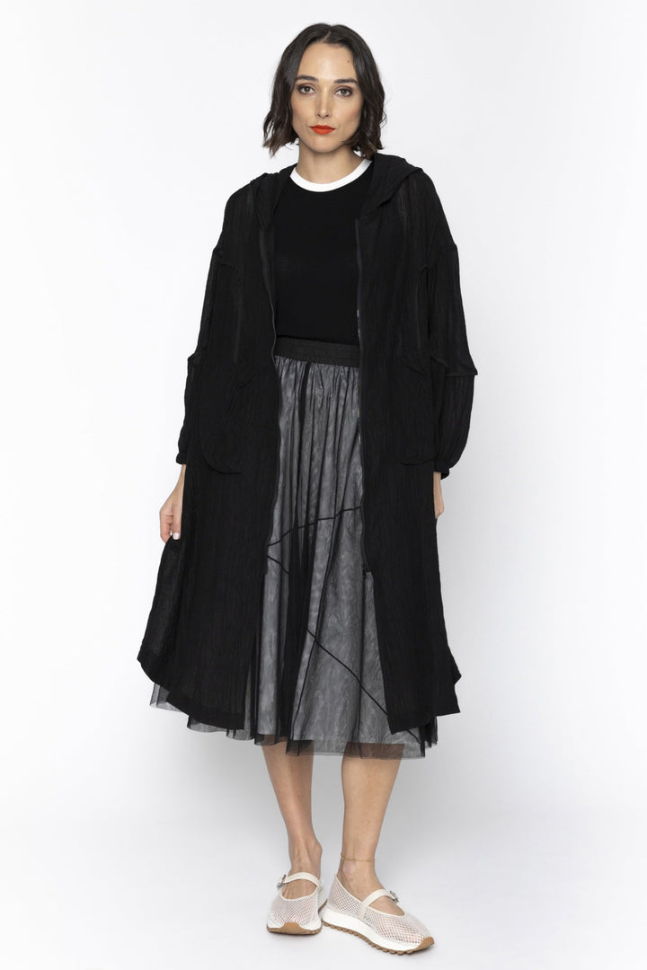 Woman wearing a black coat, black top, and sheer black skirt on a white background