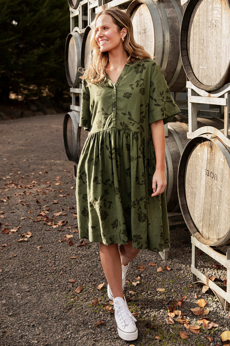 A woman standing in front of barrels, wearing a green knee-length dress with a floral pattern, a deep v neckline, and short sleeves.