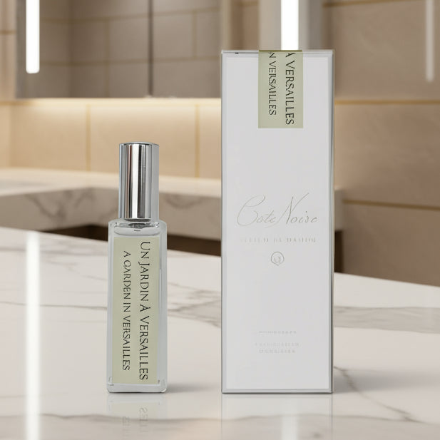 A perfume bottle labeled 'A Garden in Versailles' accompanied by its white box packaging, both featuring green text on a white background, displayed on a wooden surface with blurred objects in the background.