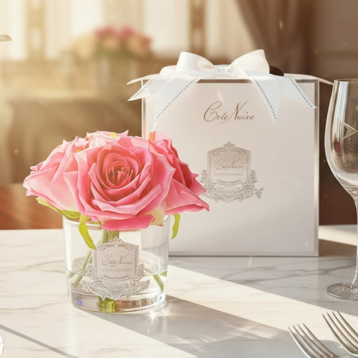 A life-like perfumed rose arranged in a clear vase with a silver badge, accompanied by a white box with a Cote Noire logo.