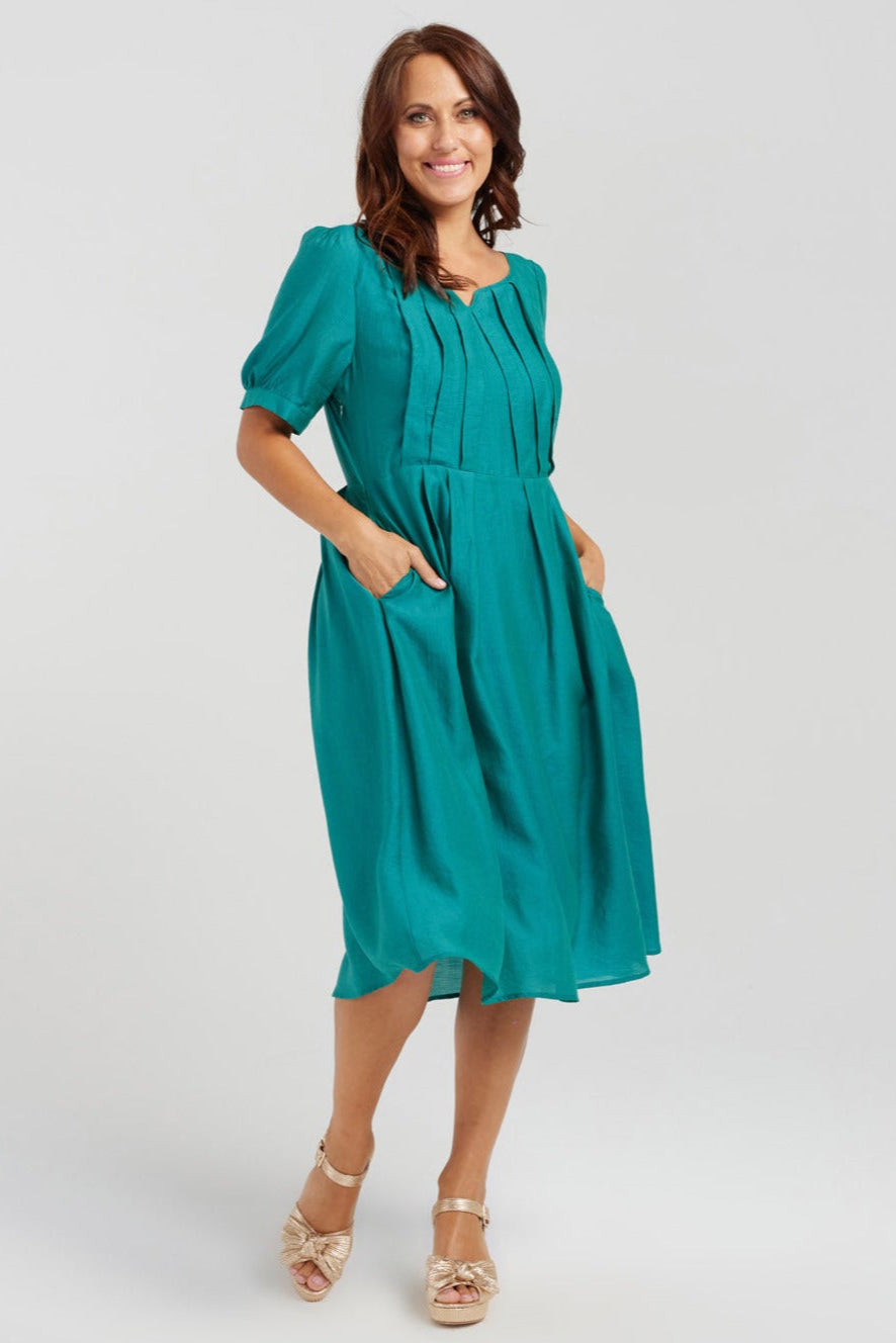 A woman standing, wearing a green dress with short sleeves and a split neckline, featuring a front bodice with bold pleats.