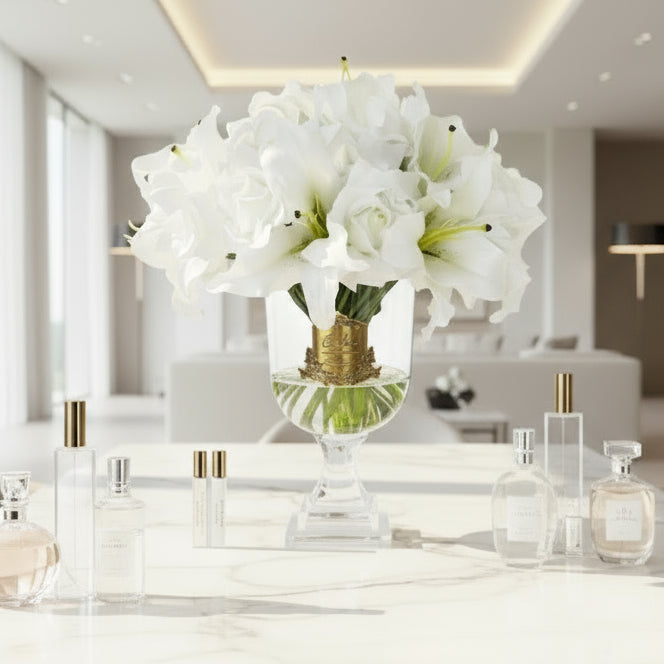 A bouquet of artificial Versailles lilies and roses in white, arranged in a luxury vase with a gold Cote Noire signature badge, accompanied by two small bottles of scented perfume.