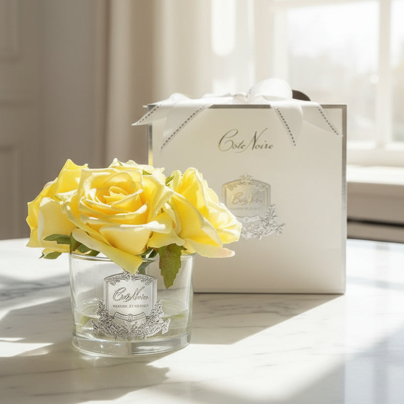 A clear glass vase with a silver signature Cote Noire badge, containing five life-like yellow perfumed roses, accompanied by two small bottles of scented perfume in a white Cote Noire ribboned box.