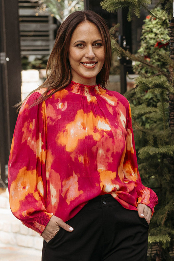 A woman wearing a vibrant multicolored blouse with a high neck and long sleeves with shirred cuffs, paired with black trousers, standing outdoors.