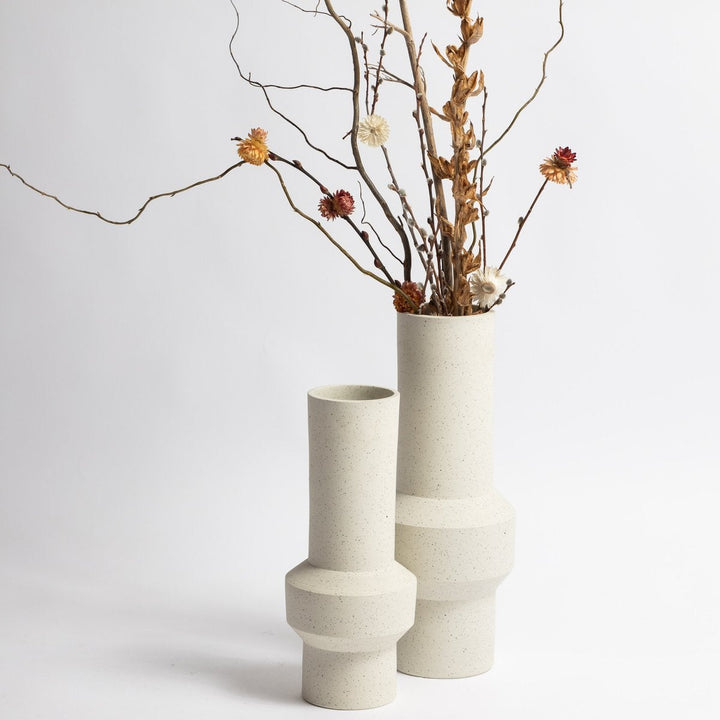 Two white ceramic vases with geometric designs, one containing dried branches and flowers on a light gray background.