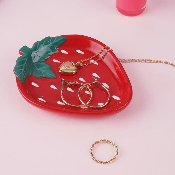 Strawberry-shaped jewelry tray with gold necklaces and rings on a pink surface.