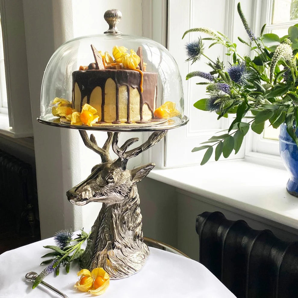 Decorative cake stand with deer head design holding a cake under a glass dome, with flowers in the background.