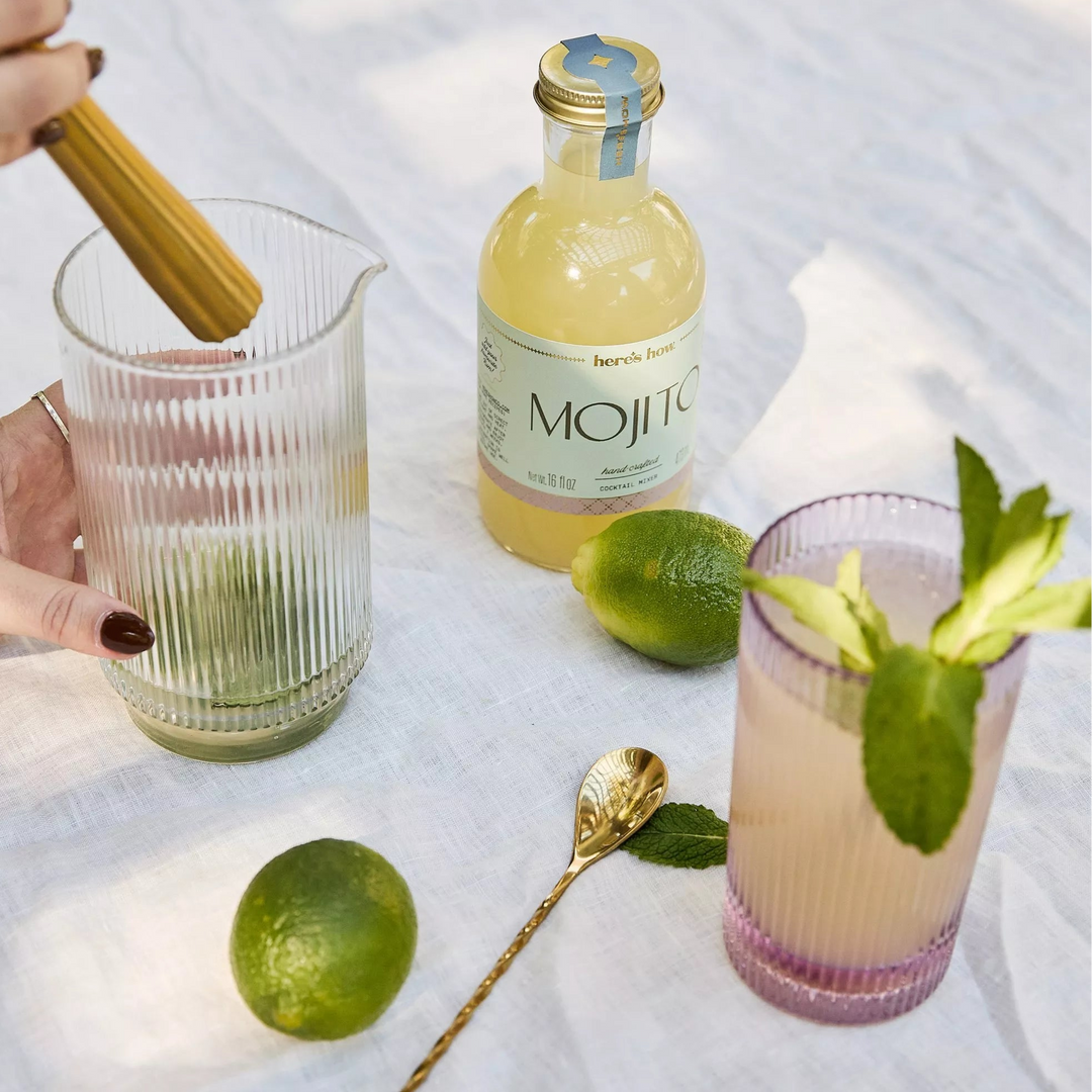 Person preparing a cocktail with lime and a bottle of mojito syrup on a white surface.
