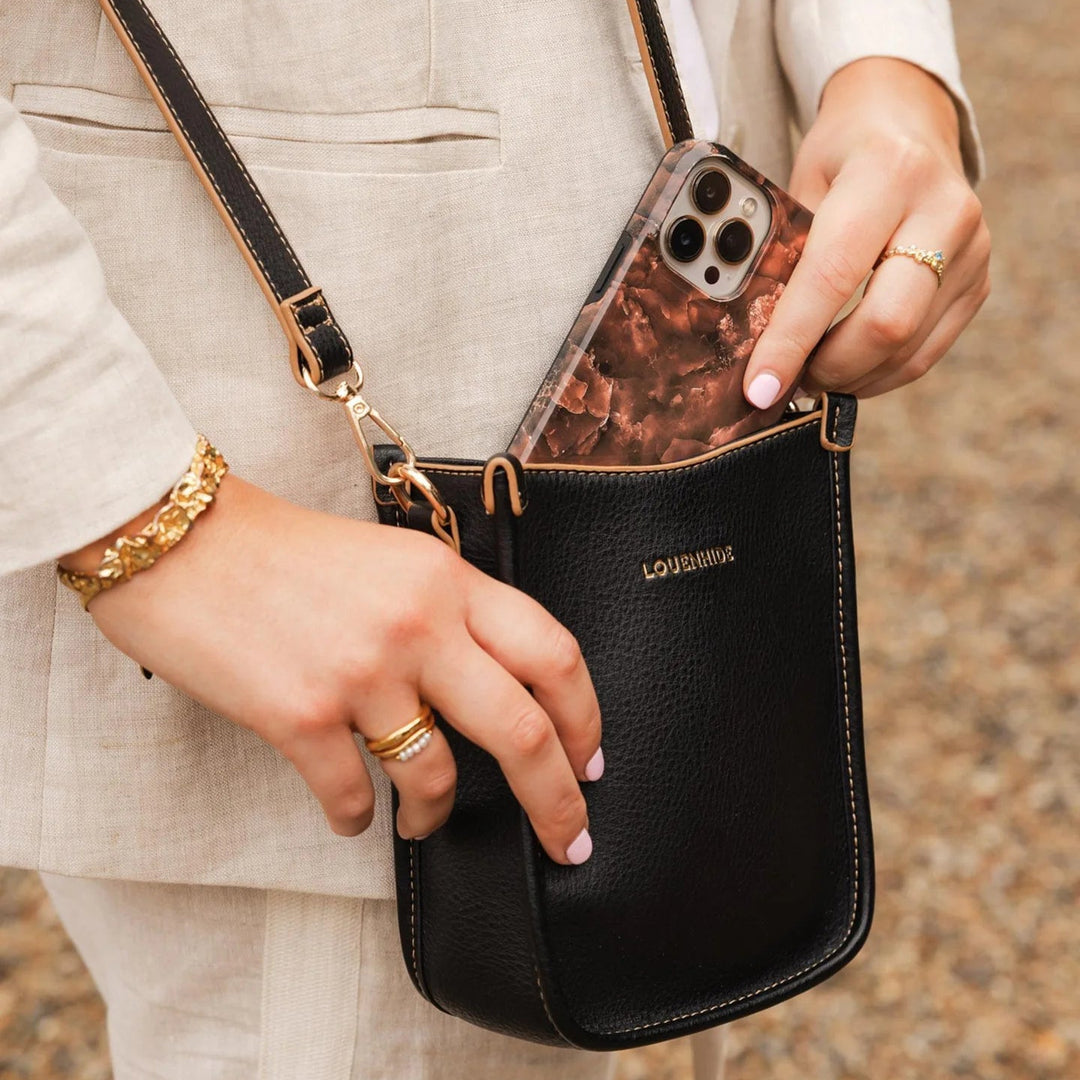 Person holding a black louenhide crossbody bag with a phone case, wearing a gold bracelet and ring.