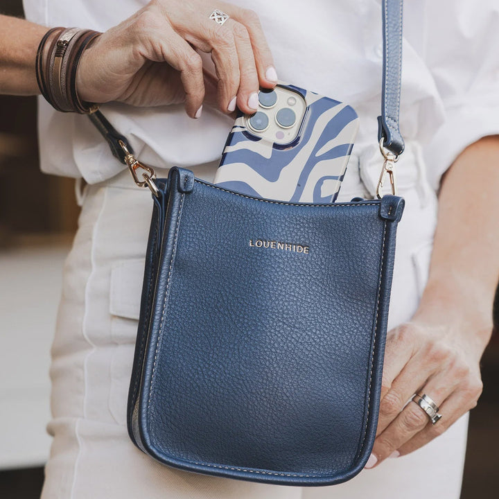 Blue crossbody louenhide parker bag with a patterned phone case held by a person wearing a white shirt.