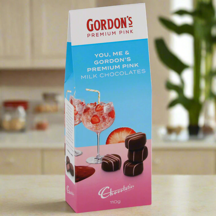 Box of Gordon's Premium Pink milk chocolates with a glass and strawberries on a pink background.