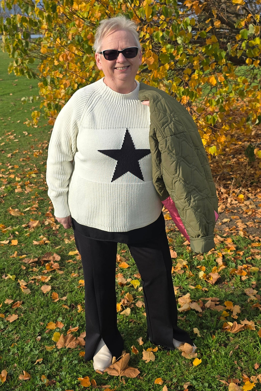 Person wearing a white sweater with a black star, dark pants, and sunglasses, standing in a grassy area with autumn leaves.