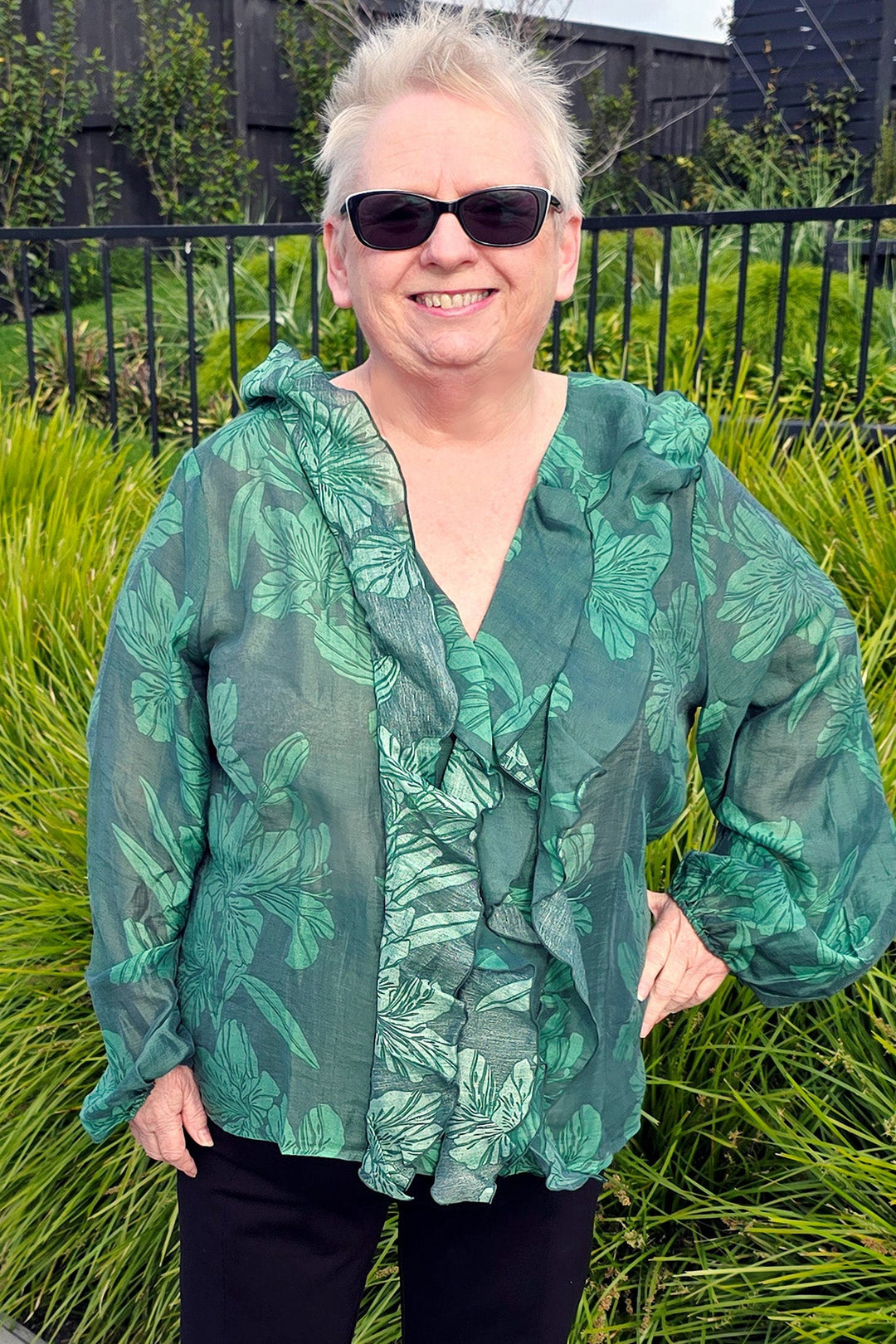 Woman wearing a green patterned blouse and black pants standing outdoors with greenery in the background.