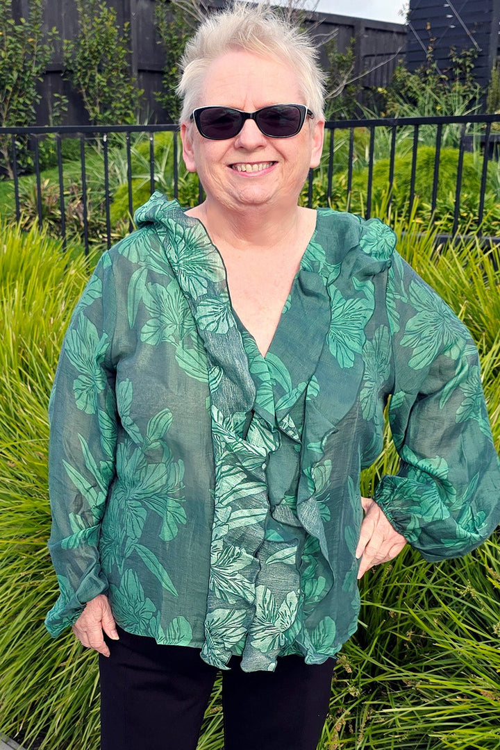 Woman wearing a green patterned blouse and black pants standing outdoors with greenery in the background.