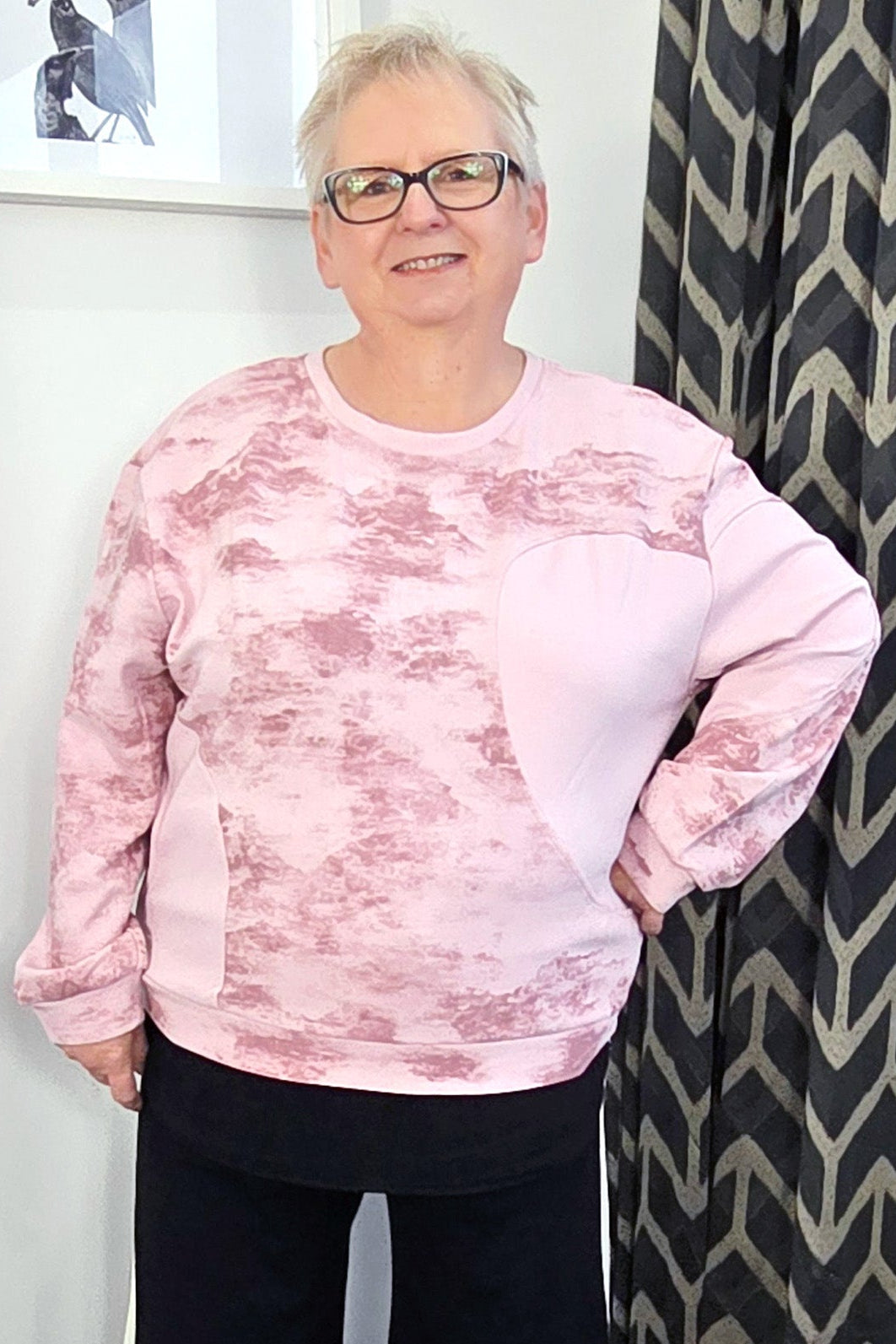 Person wearing a pink tie-dye sweatshirt and black pants standing in a room with a patterned curtain.