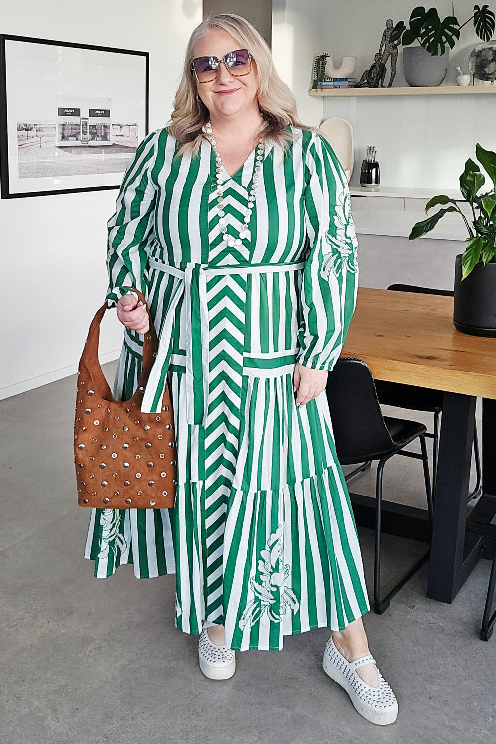 A woman wearing a green and white striped long-sleeve dress with a matching belt, holding a brown patterned handbag, standing in a modern indoor setting.