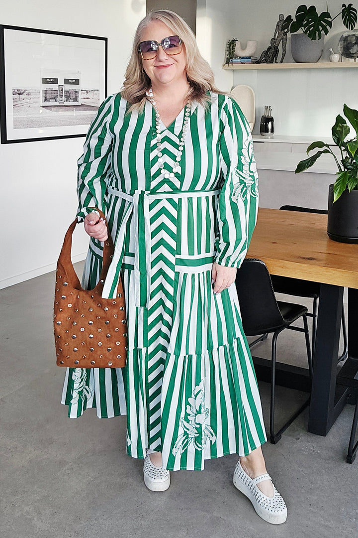 A woman wearing a green and white striped long-sleeve dress with a matching belt, holding a brown patterned handbag, standing in a modern indoor setting.