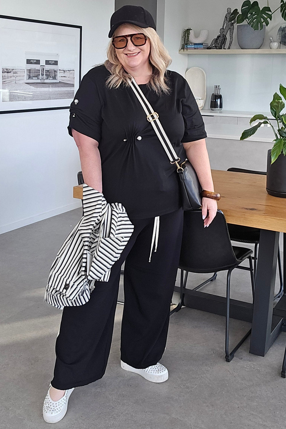 A woman posing in a modern home interior, wearing a black short-sleeve tunic top, black wide-leg trousers, a black baseball cap, and white sneakers. She is holding a striped shirt and wearing a black crossbody bag.
