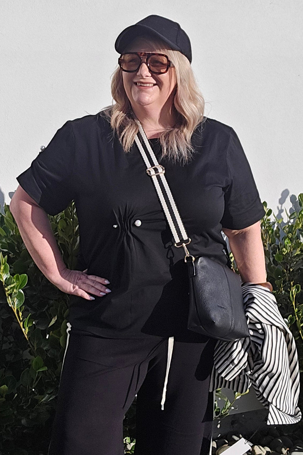 A woman wearing a black streetwear outfit with a black cap, black top, black wide-leg pants, and a black-and-white striped top underneath, posing outdoors on a paved walkway next to greenery.