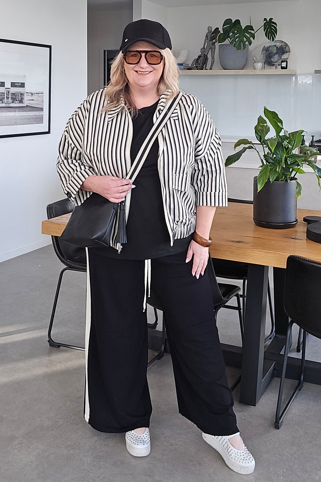 A woman posing in a modern office setting, wearing a black and white striped blazer, black top and trousers, white sneakers, and a black cap.