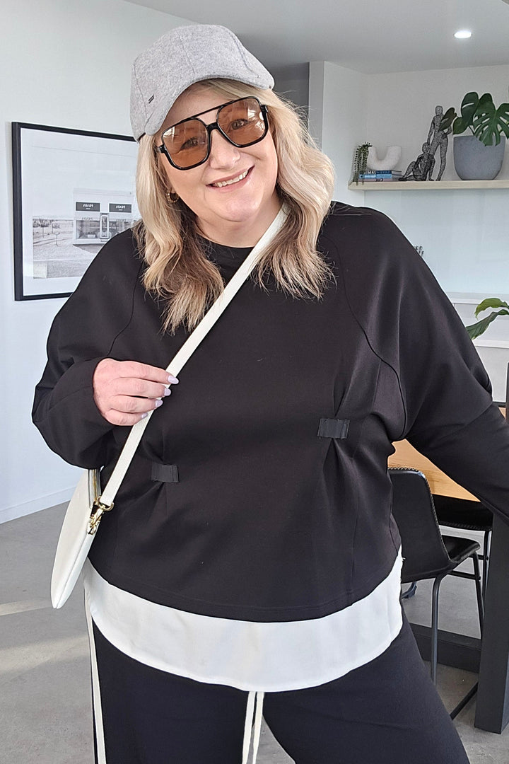 A woman wearing a black and white matching lounge set with a white drawstring bag, photographed in a modern indoor setting with a white wall and ceiling visible.