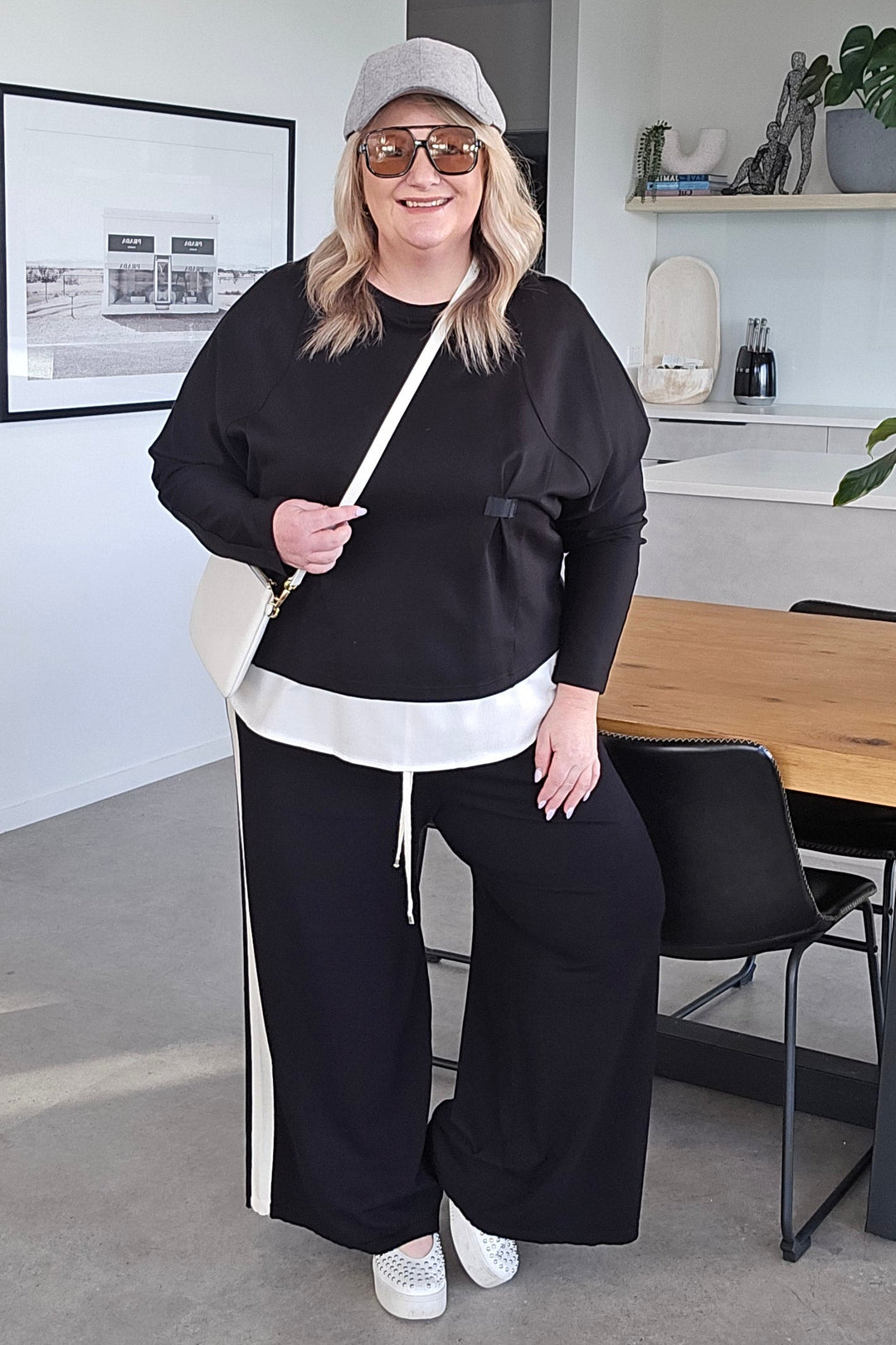 A woman wearing a black sweatshirt, black wide-leg pants with white side stripes, a white t-shirt, a white cap, and white sneakers, standing in a modern home interior.