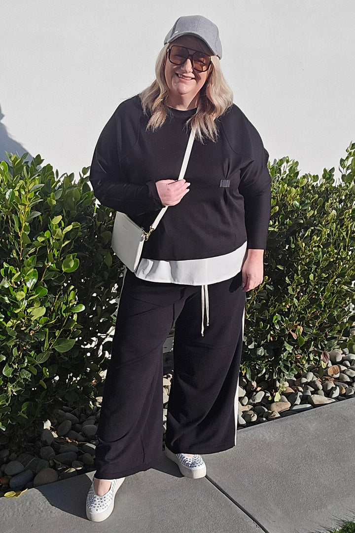 A person wearing a black tracksuit with a white waist stripe, white sneakers, and a white cap, standing outdoors on a concrete surface next to green bushes.