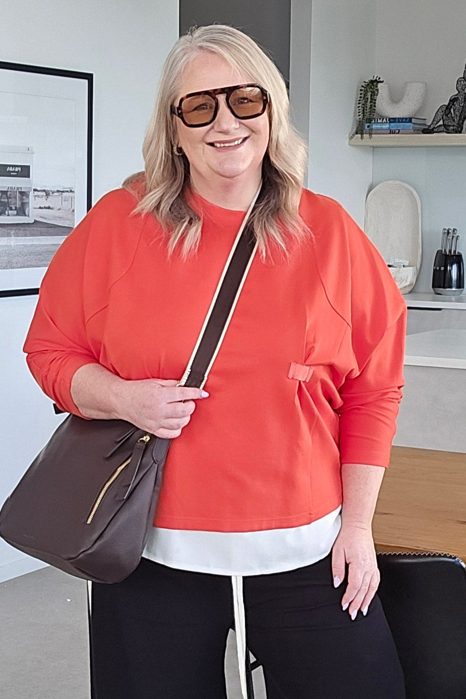 A woman wearing a coral orange long-sleeve top, black wide-leg trousers, white sneakers, and holding a brown shoulder bag, standing in a modern office space.