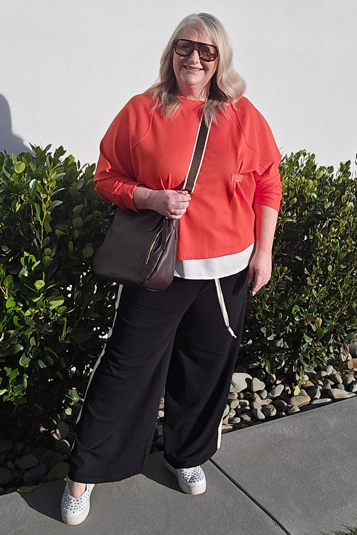 A woman wearing a bright coral-red sweatshirt, black wide-leg pants, and white sneakers stands outdoors against a white wall and green shrubs, holding a dark brown shoulder bag.