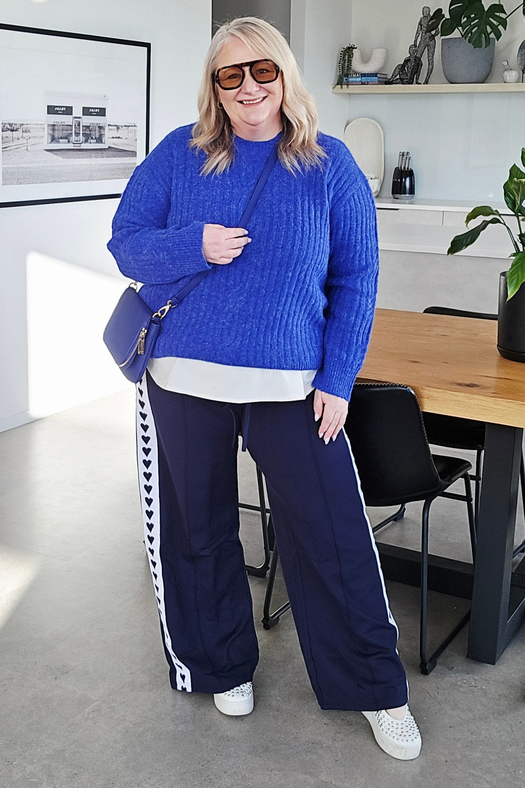 A woman wearing a bright blue knit sweater, navy wide-leg pants with white side stripes, and white sneakers, standing in a modern office interior.