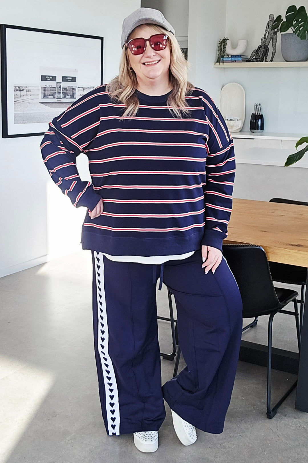A woman wearing a navy blue and pink striped long-sleeve sweatshirt and matching navy blue wide-leg sweatpants with white side stripes, posing in a modern indoor setting.