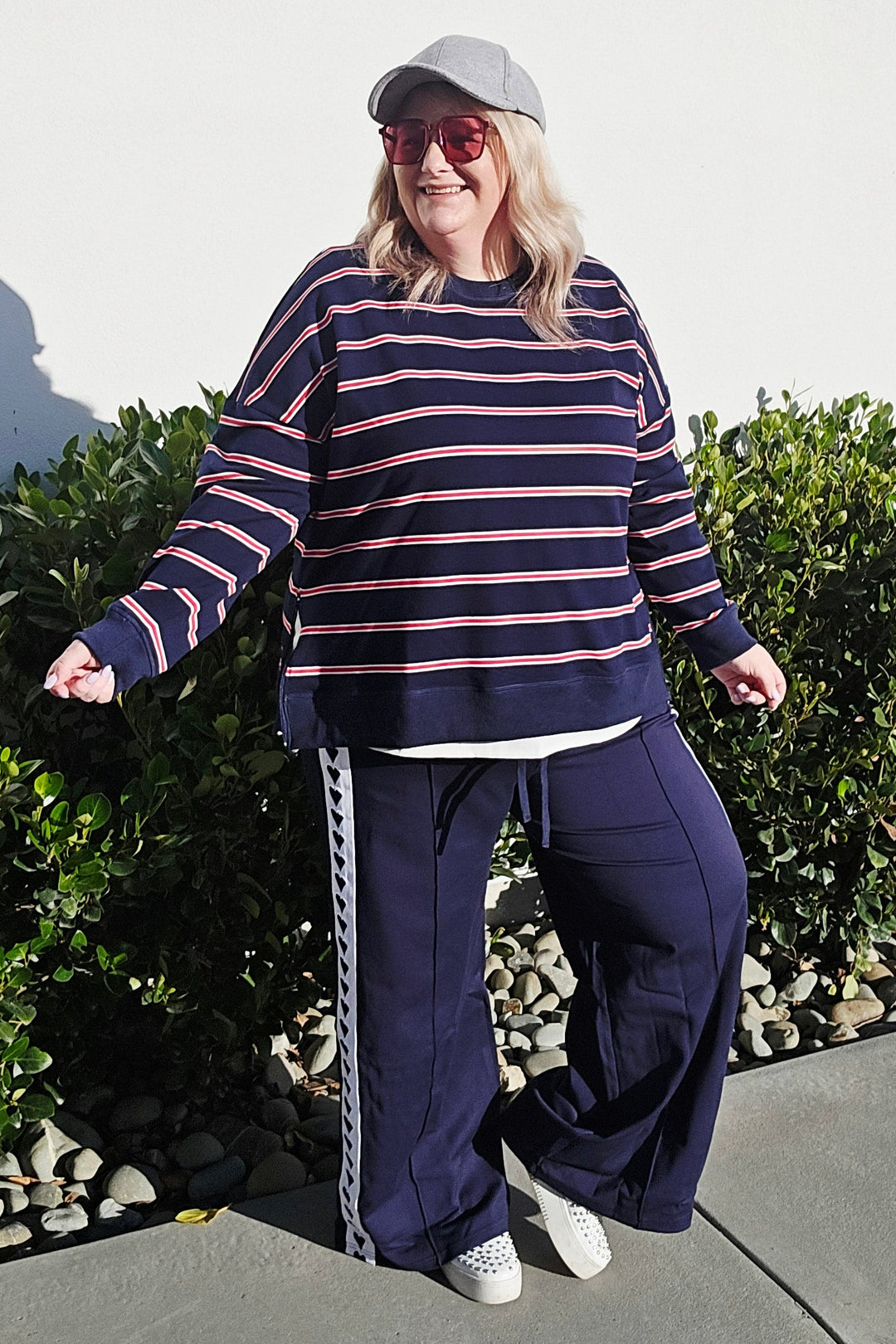A woman wearing a navy blue and pink striped long-sleeve top, matching navy wide-leg pants with a white stripe detail, a white visor, and white sneakers, standing outdoors on a concrete path next to green bushes.