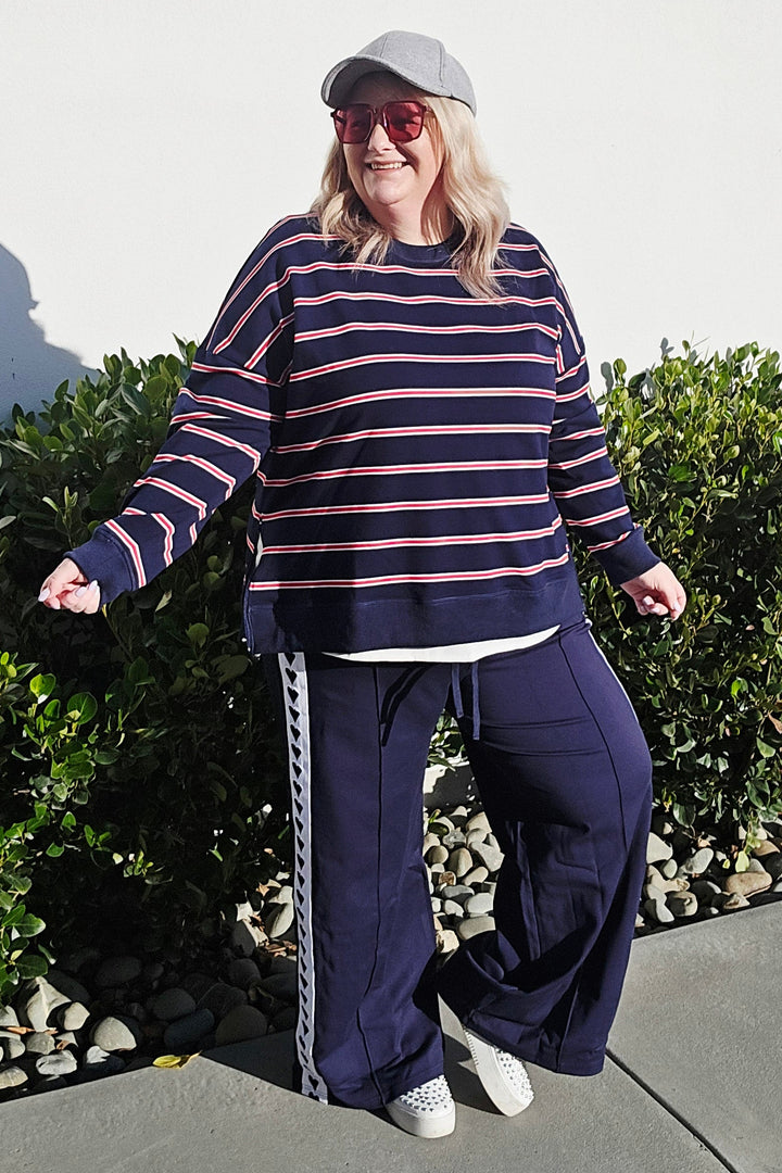 A woman wearing a navy blue and pink striped long-sleeve top, matching navy wide-leg pants with a white stripe detail, a white visor, and white sneakers, standing outdoors on a concrete path next to green bushes.