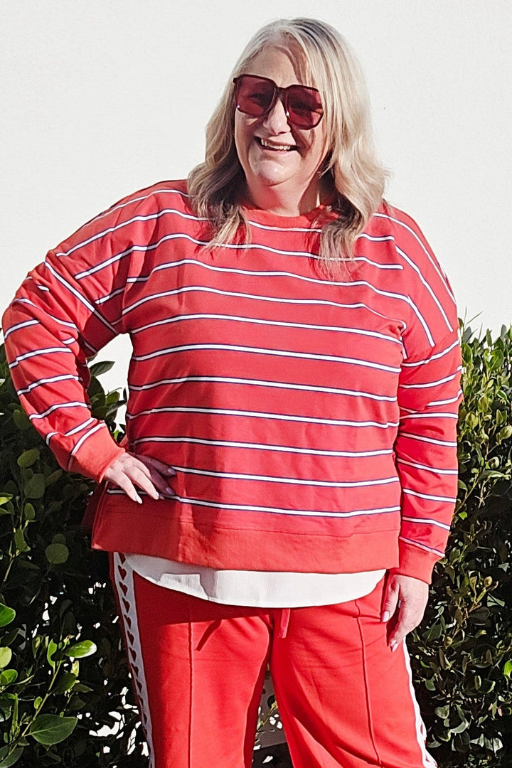 A woman wearing a matching red and white striped long-sleeve top and red pants stands outdoors on a concrete path next to green bushes.