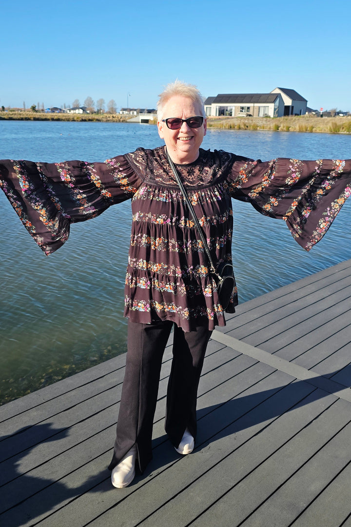 An older woman wearing a dark patterned long-sleeve top, black pants, and white shoes stands on a wooden dock with water and a building in the background.