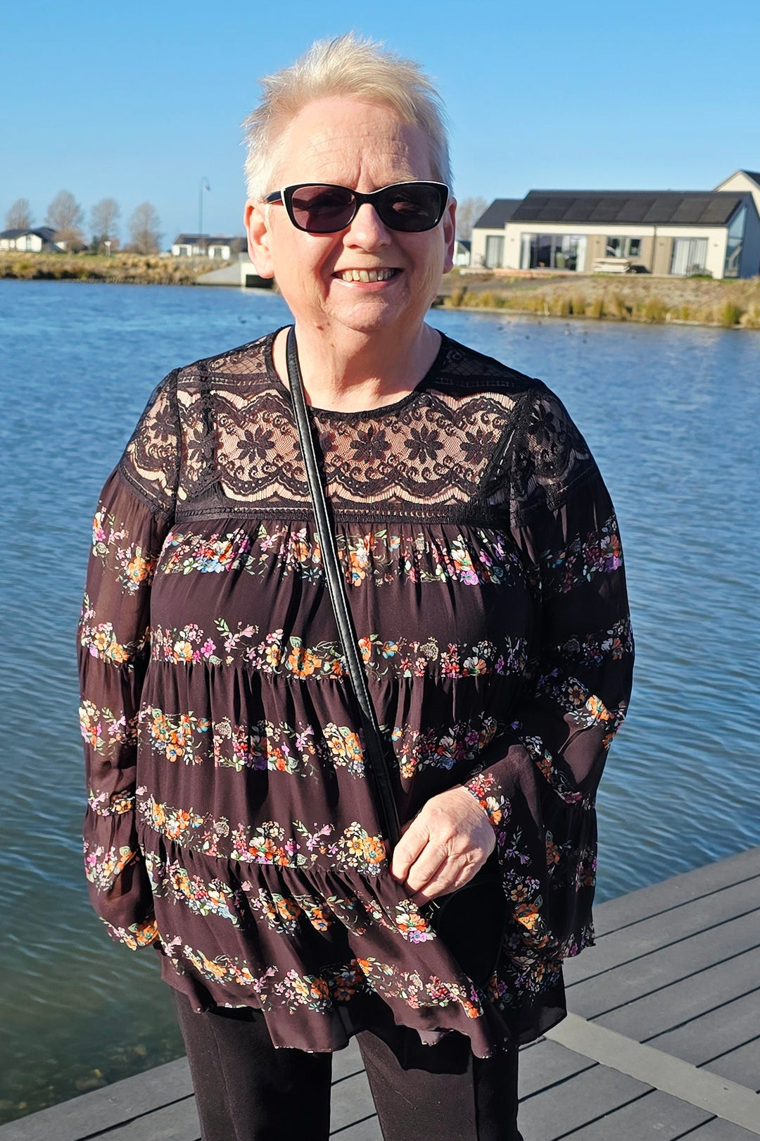 An older woman wearing a dark brown and black patterned long-sleeve top, black pants, and white shoes stands on a wooden boardwalk next to a body of water.