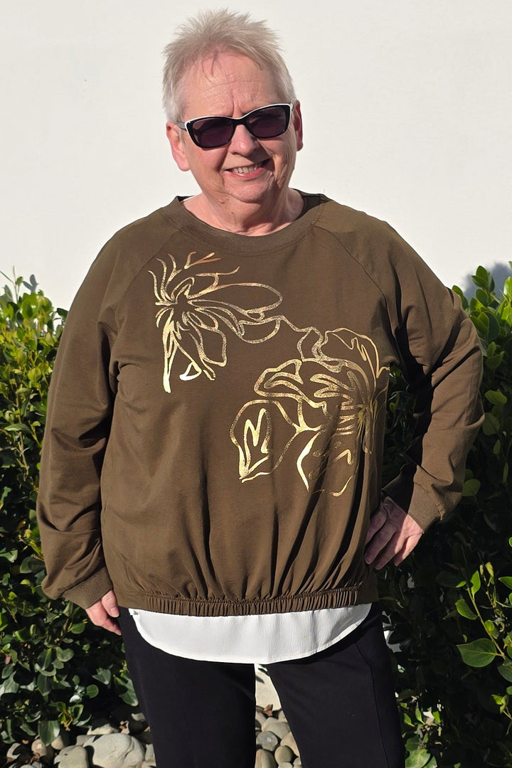 A person standing outdoors on a concrete walkway in front of a white wall and green hedge, wearing a brown sweatshirt with a gold graphic print, black pants, and white sneakers.