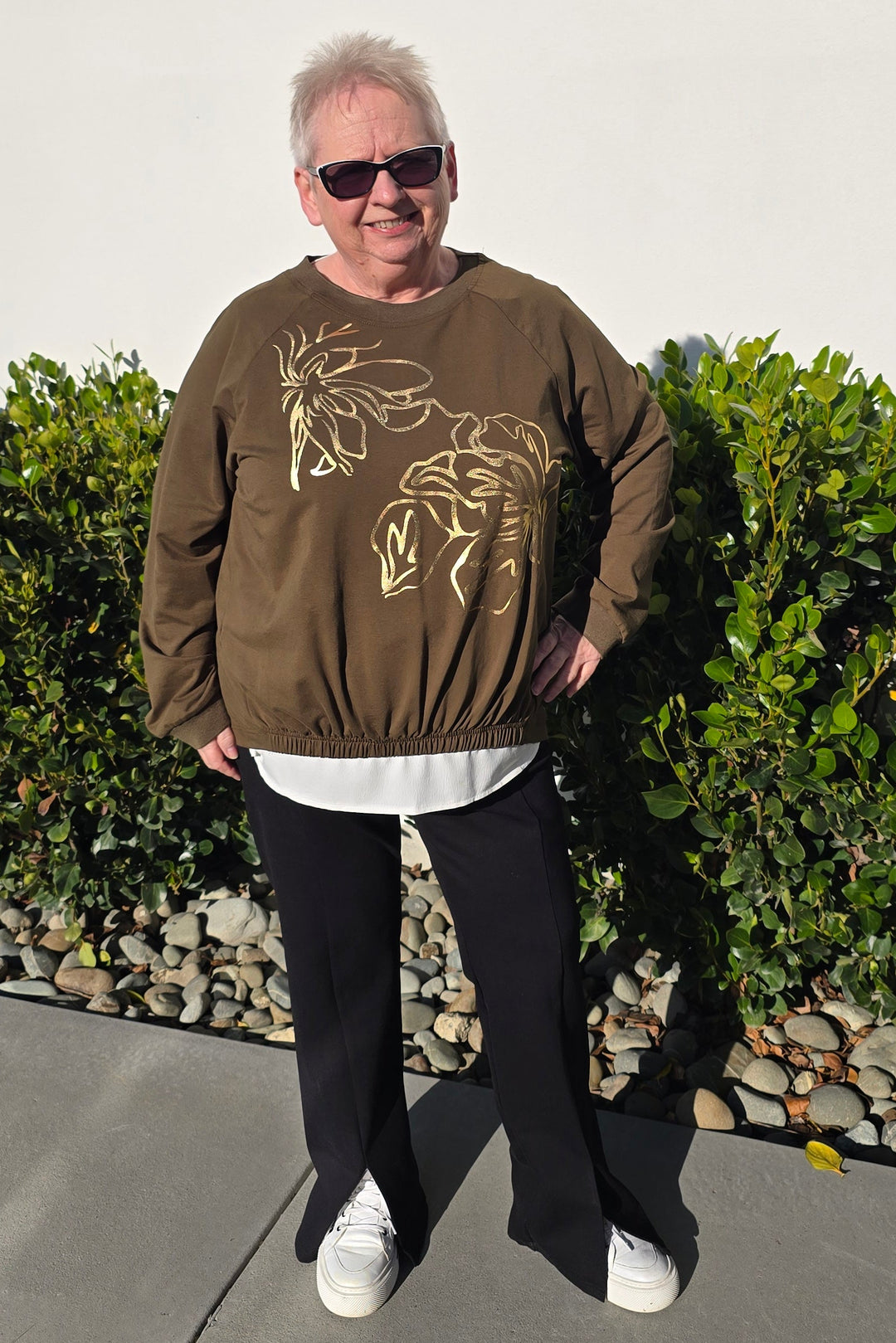 A person standing outdoors on a concrete walkway in front of a white wall and green hedge, wearing a brown sweatshirt with a gold graphic print, black pants, and white sneakers.