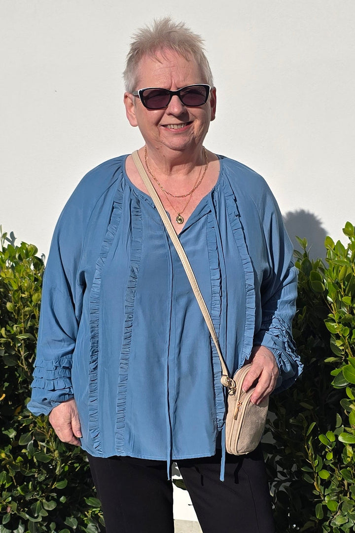 An older woman wearing a blue button-up shirt, black pants, and beige shoes stands outdoors on a paved path next to green bushes, holding a small beige handbag.