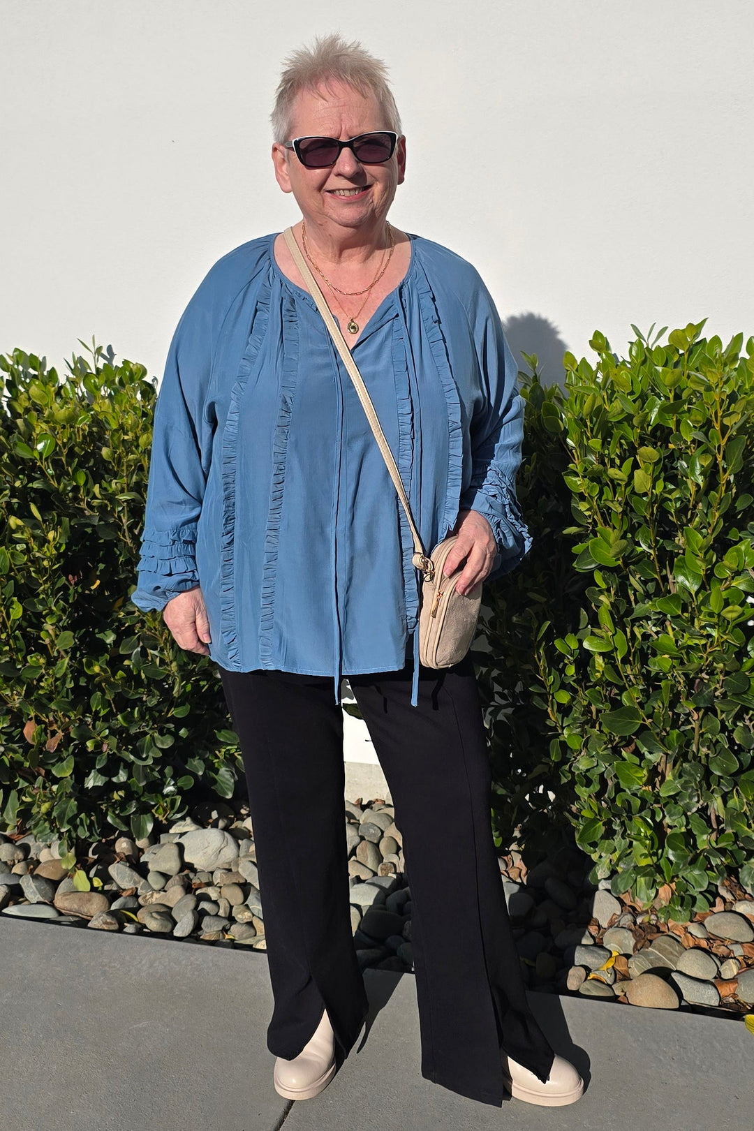 An older woman wearing a blue button-up shirt, black pants, and beige shoes stands outdoors on a paved path next to green bushes, holding a small beige handbag.