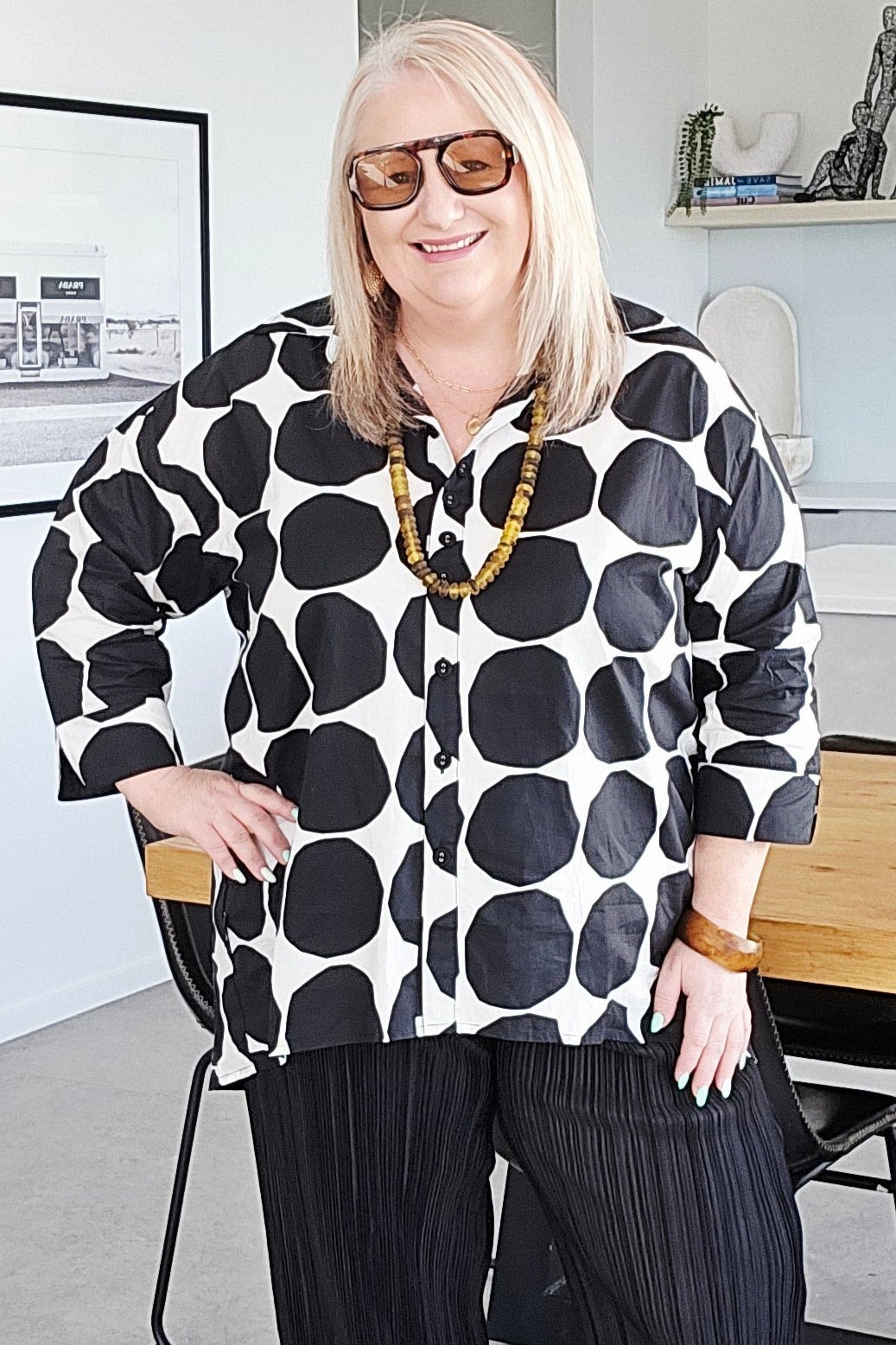 A woman wearing a black and white polka dot shirt, black pleated trousers, and white sneakers stands in a modern home interior.