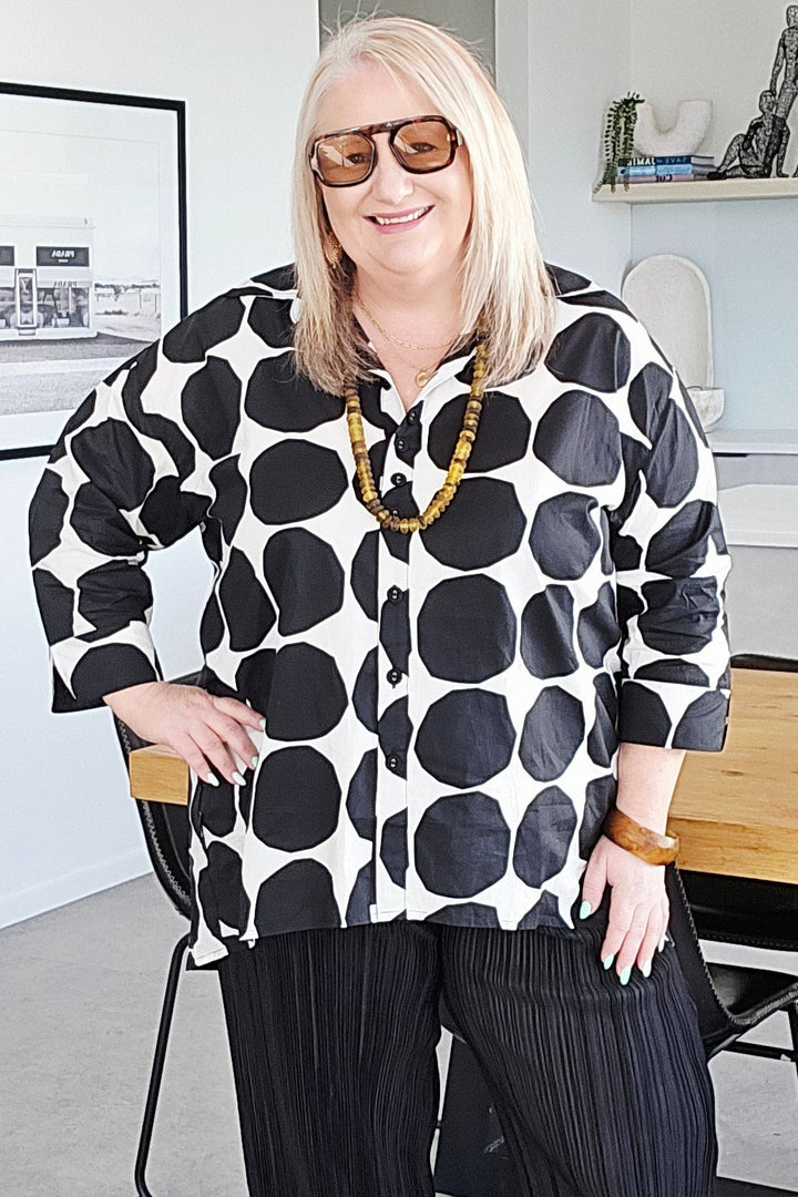 A woman wearing a black and white polka dot shirt, black pleated trousers, and white sneakers stands in a modern home interior.