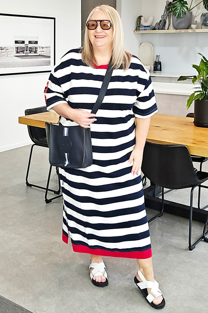A woman wearing a black and white striped midi dress with red trim, sunglasses, and white slide sandals, carrying a black shoulder bag in a modern home interior setting.