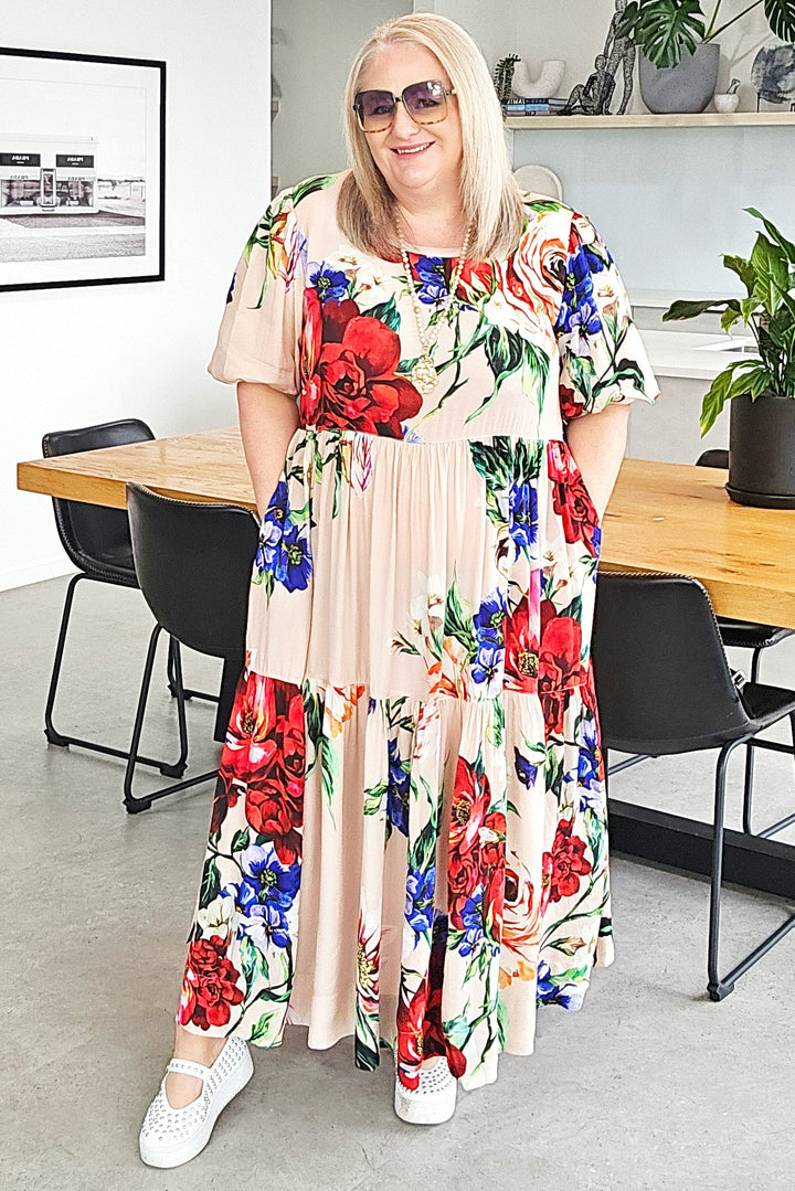 A woman wearing a beige maxi dress with a vibrant red, blue, and green floral print stands in a modern dining room setting.