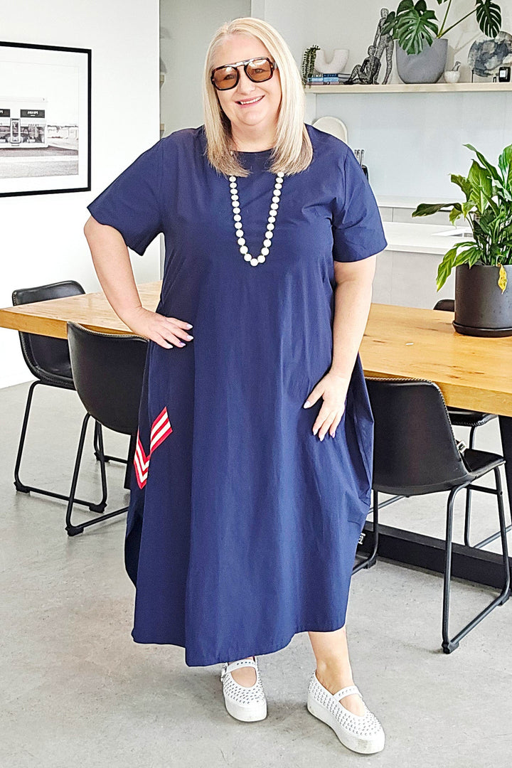 A woman wearing a navy blue midi dress with a tie-neck detail, sunglasses, and a pearl necklace stands in a modern office or cafe setting with a wooden table, chairs, and plants in the background.