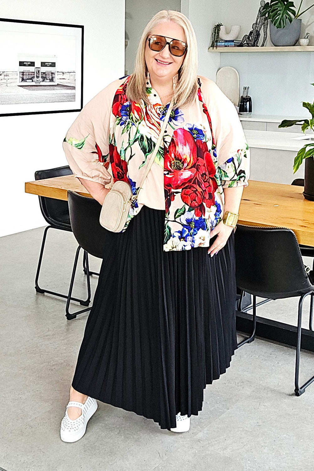 A woman modeling a stylish outfit consisting of a beige bomber jacket with a vibrant red, blue, and green floral print, a long black pleated skirt, and white sneakers, standing in a modern minimalist room.