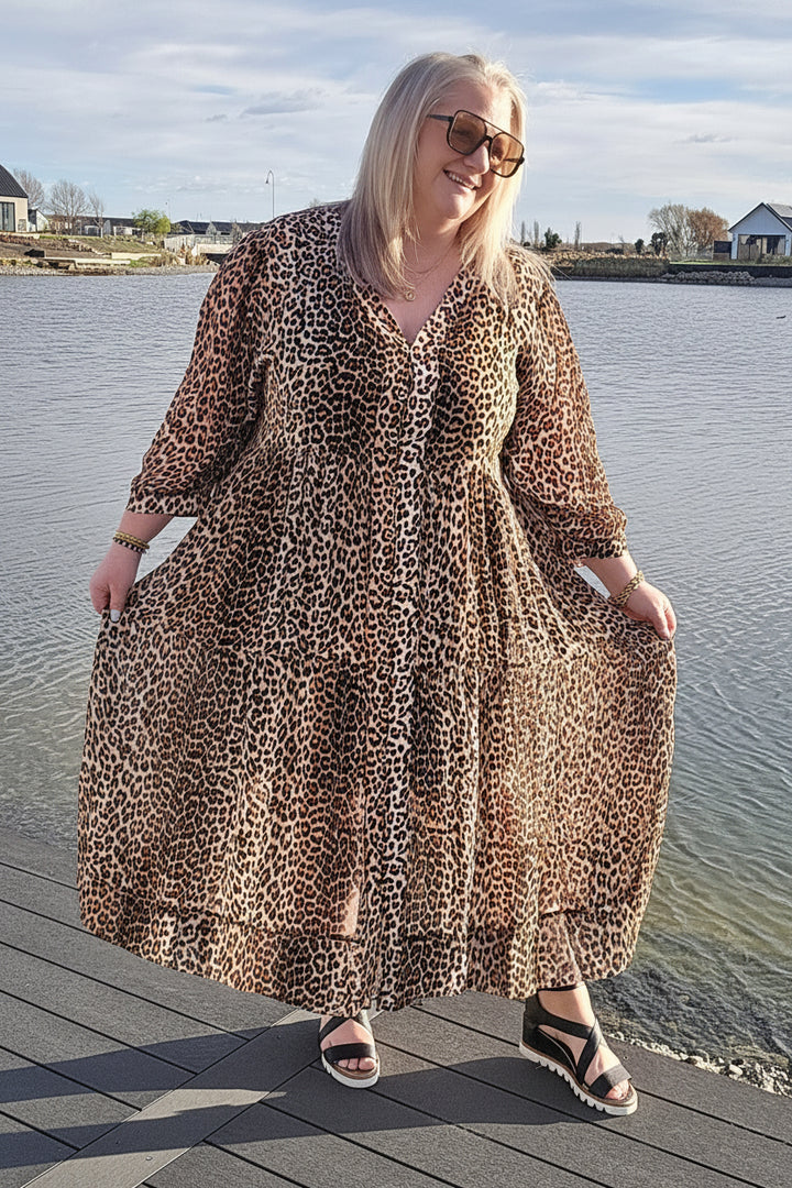 A woman wearing a leopard print maxi dress and sunglasses stands on a wooden dock next to a lake, holding out the sides of the dress to show its flow.