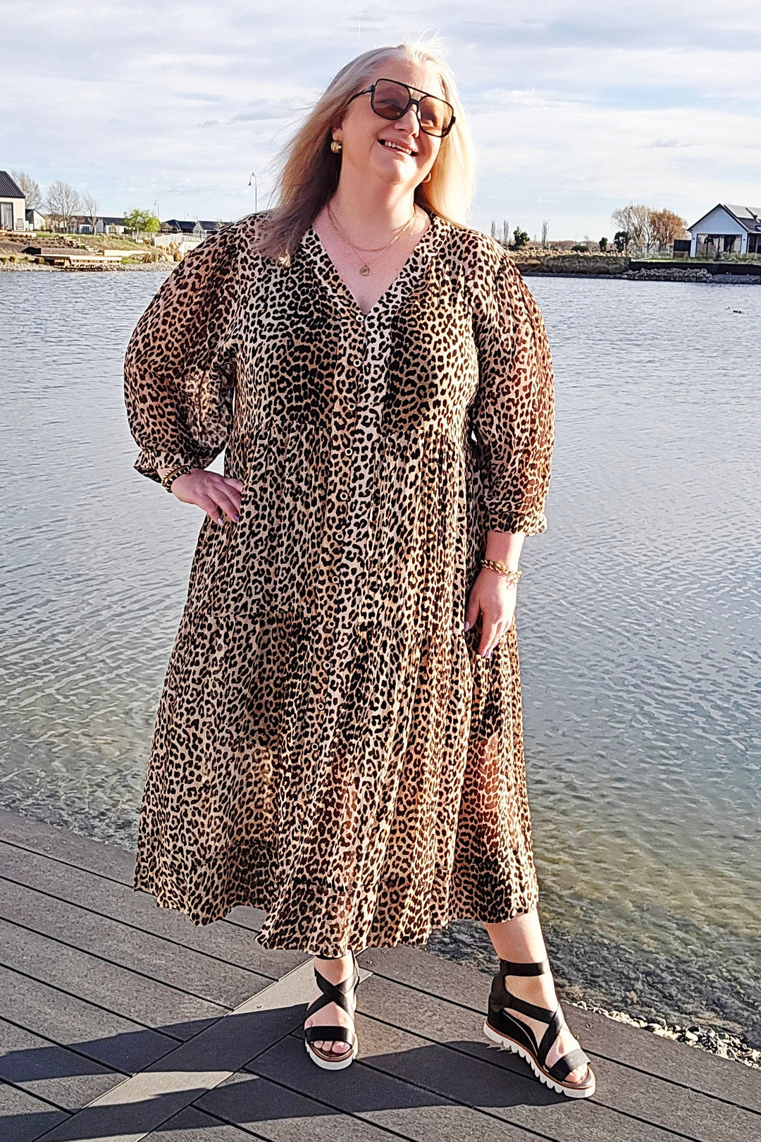 A woman wearing a leopard-print midi dress and black strappy sandals stands on a wooden dock next to a calm lake under a cloudy sky.