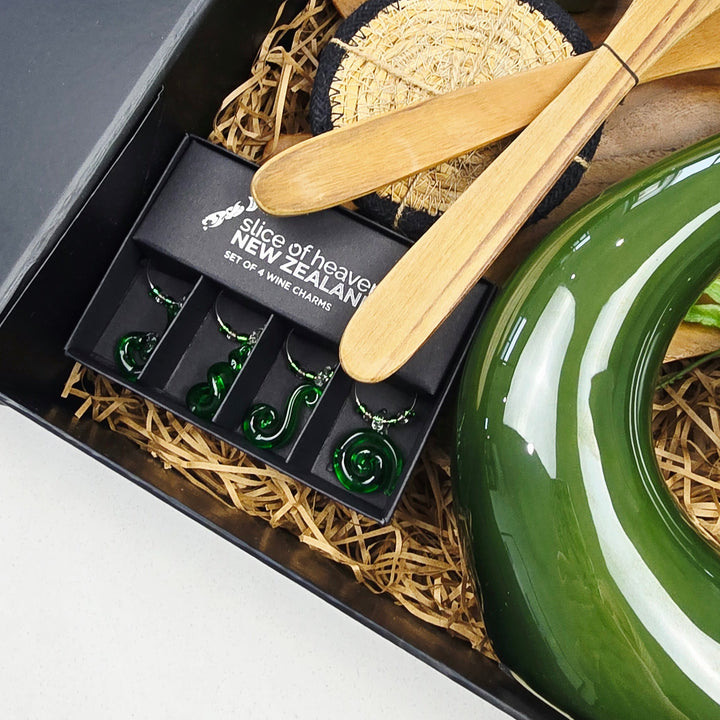 A curated gift set in a black box with natural straw filler, featuring a glossy green ceramic bowl, wooden utensils, a carved green New Zealand tiki figure, and a black jewelry tray with green earrings.