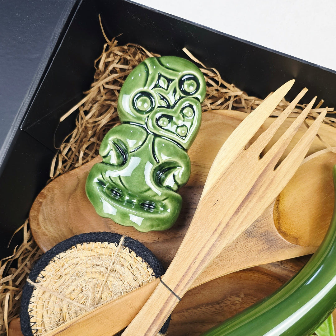 A Māori-inspired gift set in a black box with straw lining, featuring a green carved skull token, a wooden spoon, a wooden fork with a wooden handle, a green curved decorative piece, and a small woven basket.